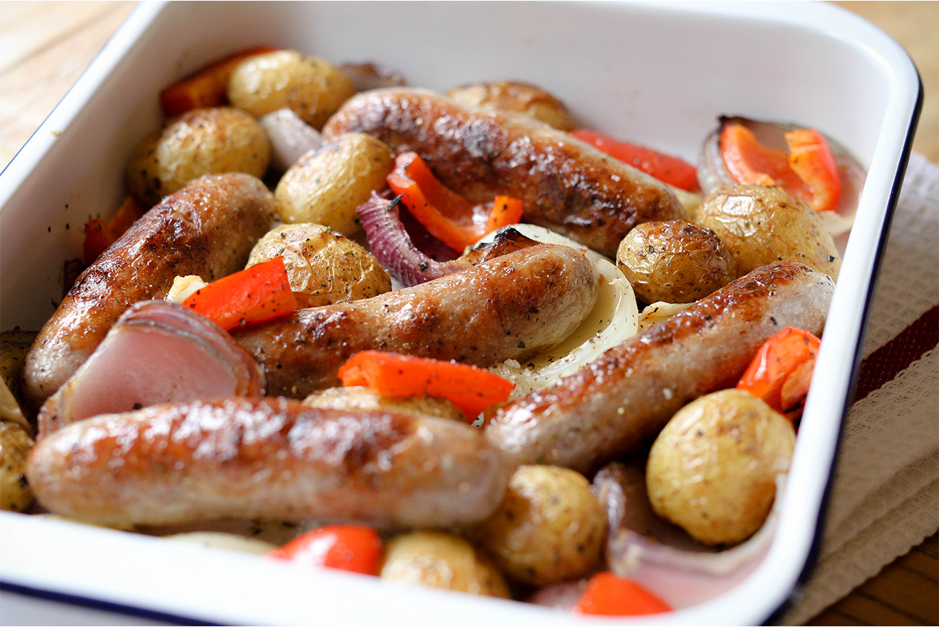 An enamel tray filled with oven cooked sausages, new potatoes, red peppers and onions